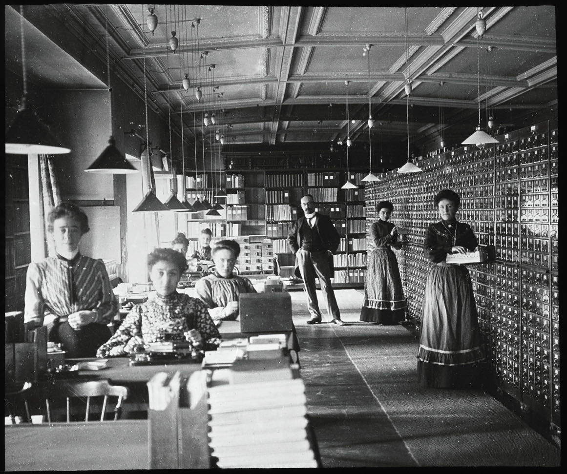 Staff of the International Institute of Bibliography seated at long tables, writing and classifying index cards beside walls of card catalog drawers, around 1900